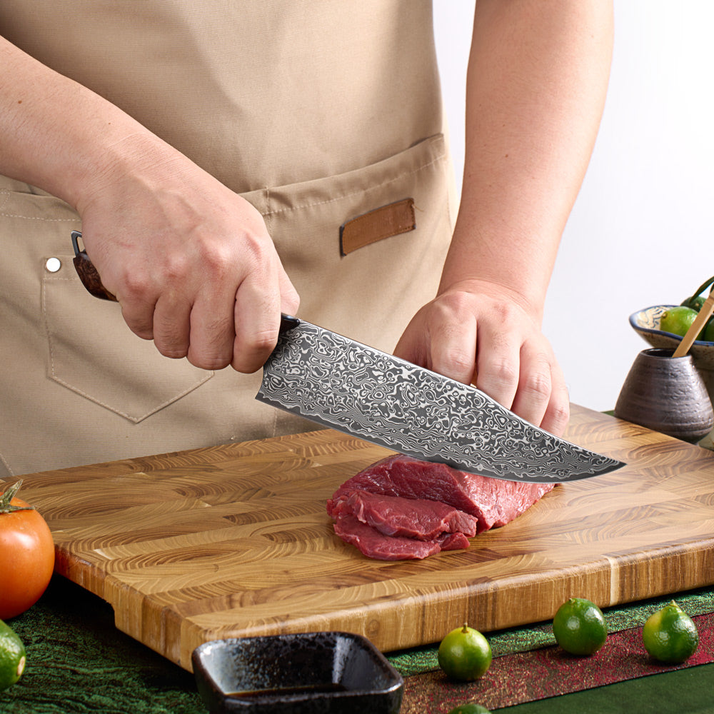 A close-up of a person in a beige apron holding a large kitchen knife with a patterned steel blade, slicing raw red meat on a wooden cutting board. Small green limes and dark bowls are visible around the board.