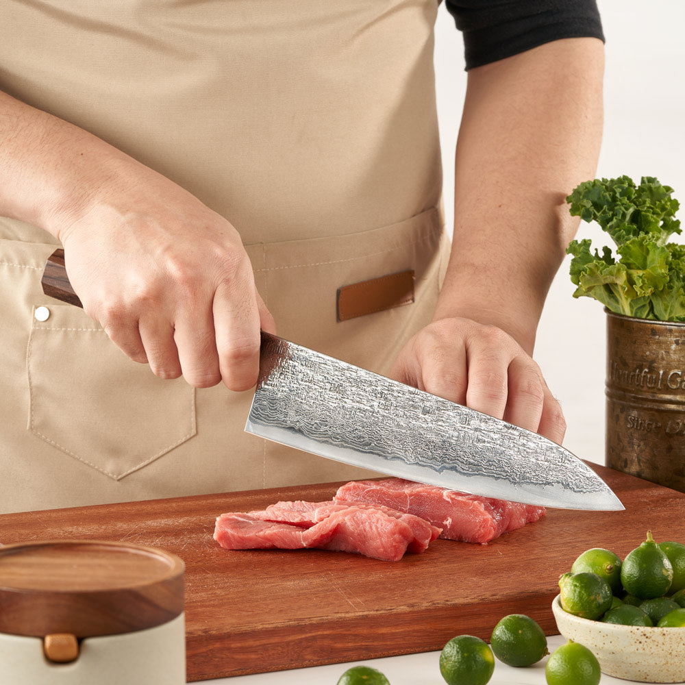 Person cutting meat on a wooden board with a chef's knife, surrounded by limes and greens.