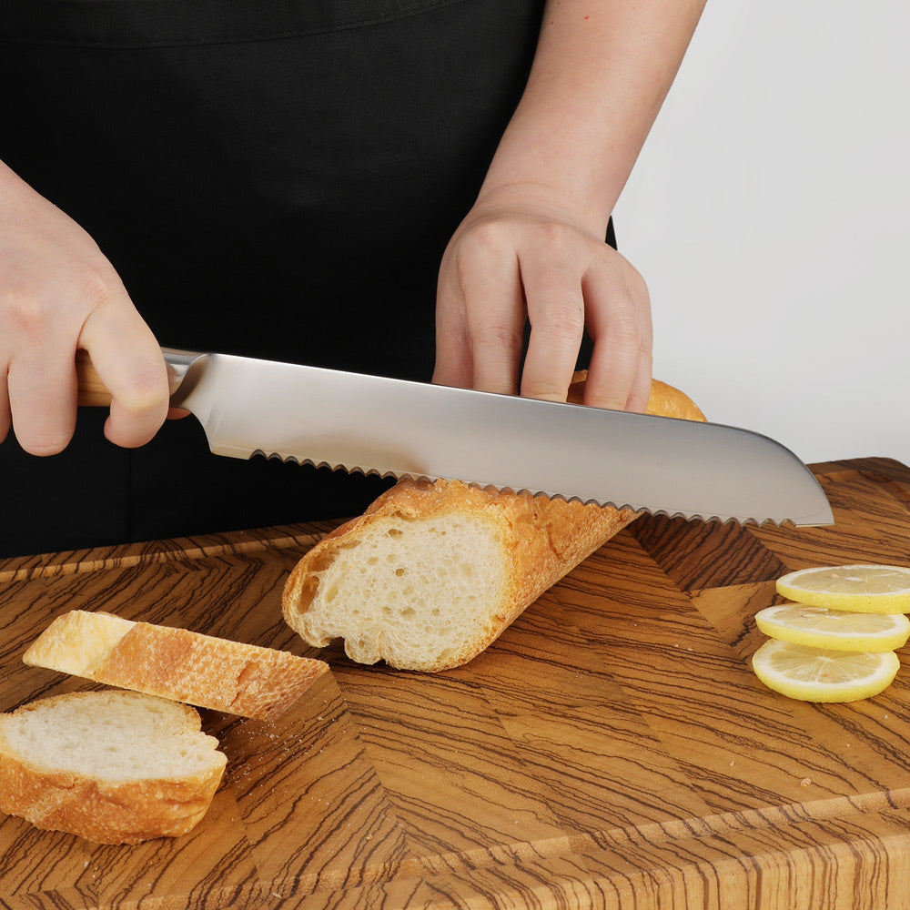A serrated bread knife with a silver blade and light wood handle being used to slice a loaf of French bread on a wooden cutting board, with lemon slices nearby.