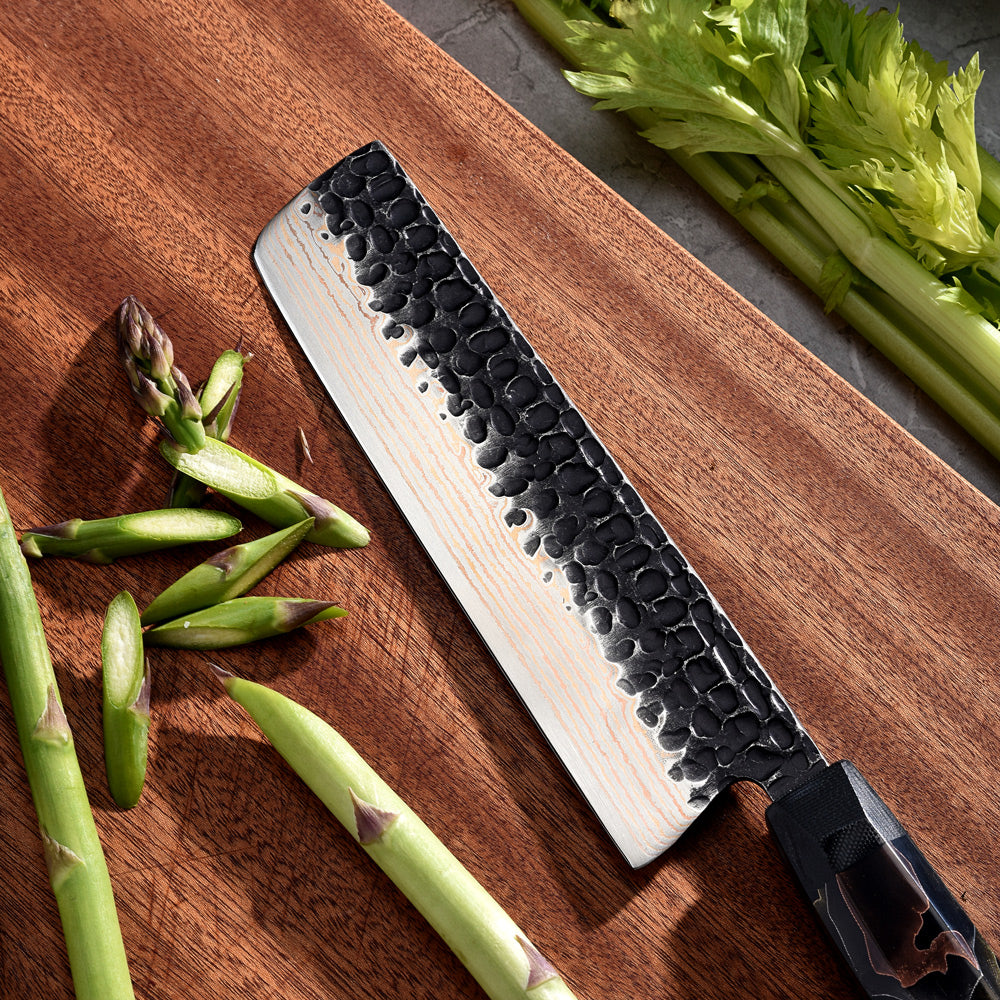 Professional Kitchen knife with black damascus hammered blade on a wooden cutting board with asparagus and celery.